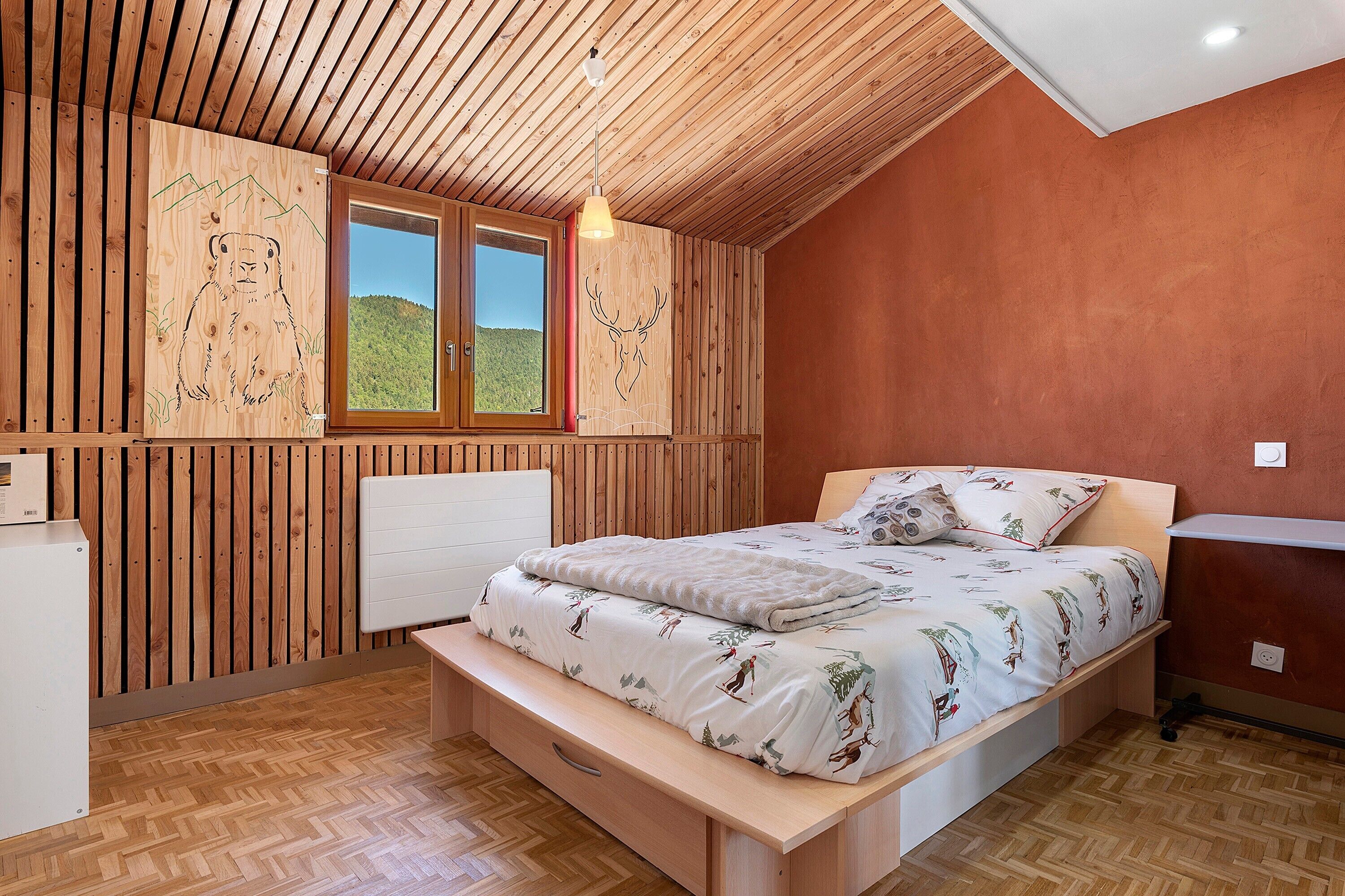 A bedroom with a wooden slatted ceiling, a bed with a patterned duvet, a window with a view of a green landscape, and a radiator.
