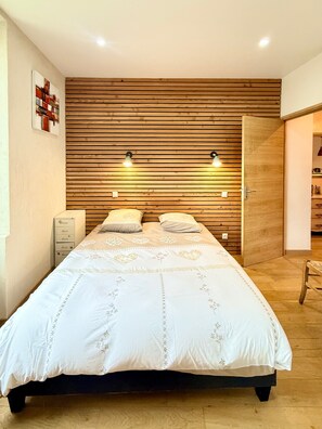 A bedroom with a wooden slat wall, a bed with a white bedspread, two bedside lamps, and a nightstand with drawers. - L’Authentique du Vercors – Spacious Modern Wood-Style Apartment with Mountain Views (Villard-de-Lans)