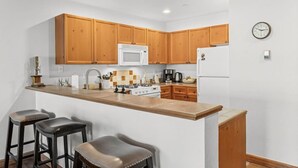 Une cuisine avec des meubles de rangement en bois, un micro-ondes blanc, une cuisinière, un réfrigérateur, une horloge et trois tabourets de bar.