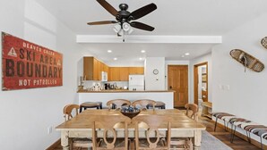 Un coin repas avec une table et des chaises en bois, un ventilateur de plafond et une cuisine en arrière-plan.