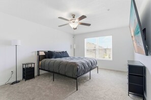 Ein Schlafzimmer mit Bett, Deckenventilator, Nachttisch, Kommode und Fenster mit Jalousien.