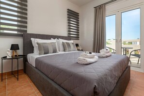 A hotel room with a large bed, a small table with a lamp, a nightstand, and a view of the outdoors. - Villa Avia Pefkos by Villa Plus (Pefki)