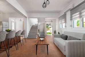 A modern living room with a sofa, a dining area with chairs, a staircase, and an air conditioning unit. - Villa Avia Pefkos by Villa Plus (Pefki)