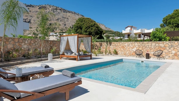 Un área de piscina con terraza de madera, sillas de descanso, un muro de piedra y montañas al fondo.