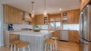 Une cuisine moderne avec des meubles en bois, un réfrigérateur en acier inoxydable, un îlot central avec des tabourets, un éclairage suspendu et une vue sur l’extérieur.