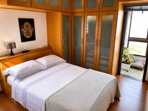 A bedroom with a bed, wooden wardrobe, and a window with a view. - Panoramic view of the ocean, lagoon, and mountains - Barra da Tijuca Beach (Rio de Janeiro)