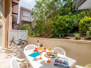 Uma mesa posta com garrafa, copos e cardápio, rodeada por cadeiras, com uma bicicleta e vegetação ao fundo.