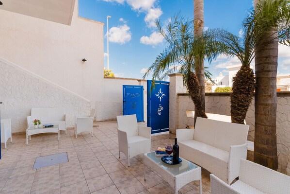 A sunlit outdoor seating area with white wicker furniture, a blue door, and a palm tree. - B&B Gioia del Mare - Camera Deluxe PP (Torre Lapillo)