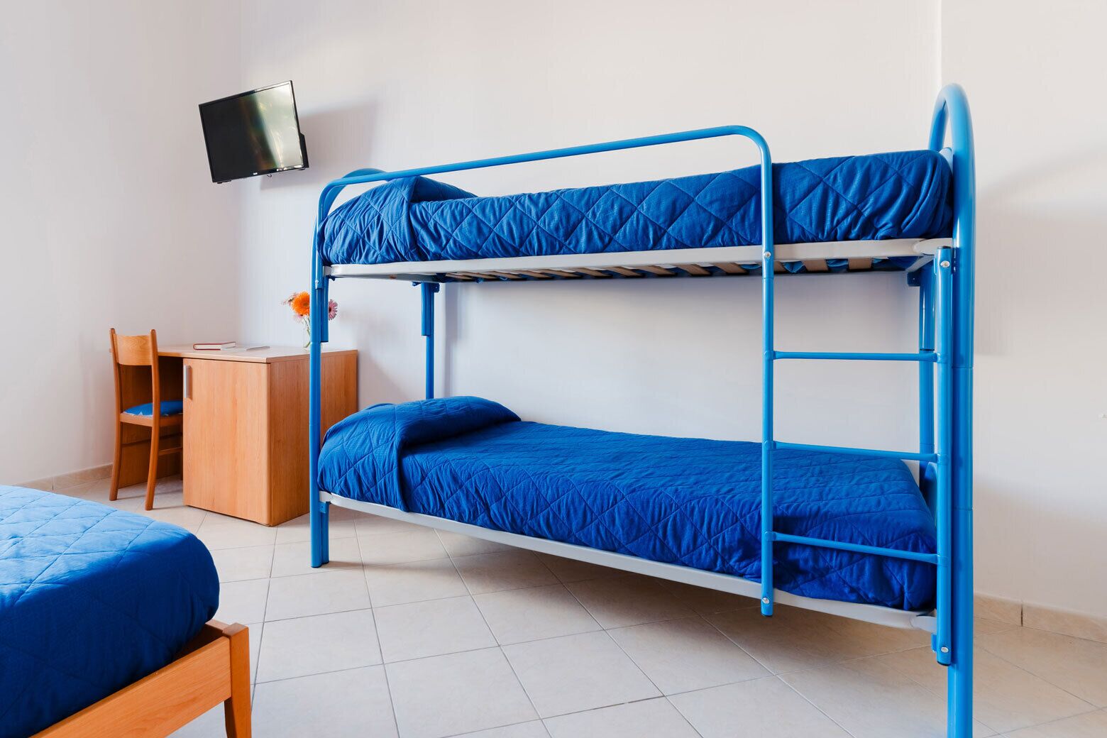 Ein Stockbett mit blauer Bettwäsche, ein Schreibtisch mit Stuhl und ein Fernseher an der Wand.