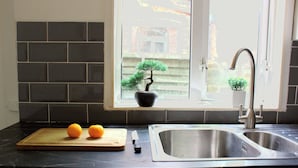A kitchen with a black countertop, a wooden cutting board with two oranges, a knife, a sink, and a window with a view of a building and a tree. - 3-Bed Bearwood | Midland Met, No CAZ, Free Parking (Smethwick)