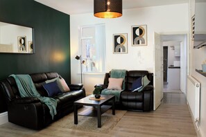 Un salon comprenant un canapé, un fauteuil et une table basse. Une porte donne accès à la cuisine.