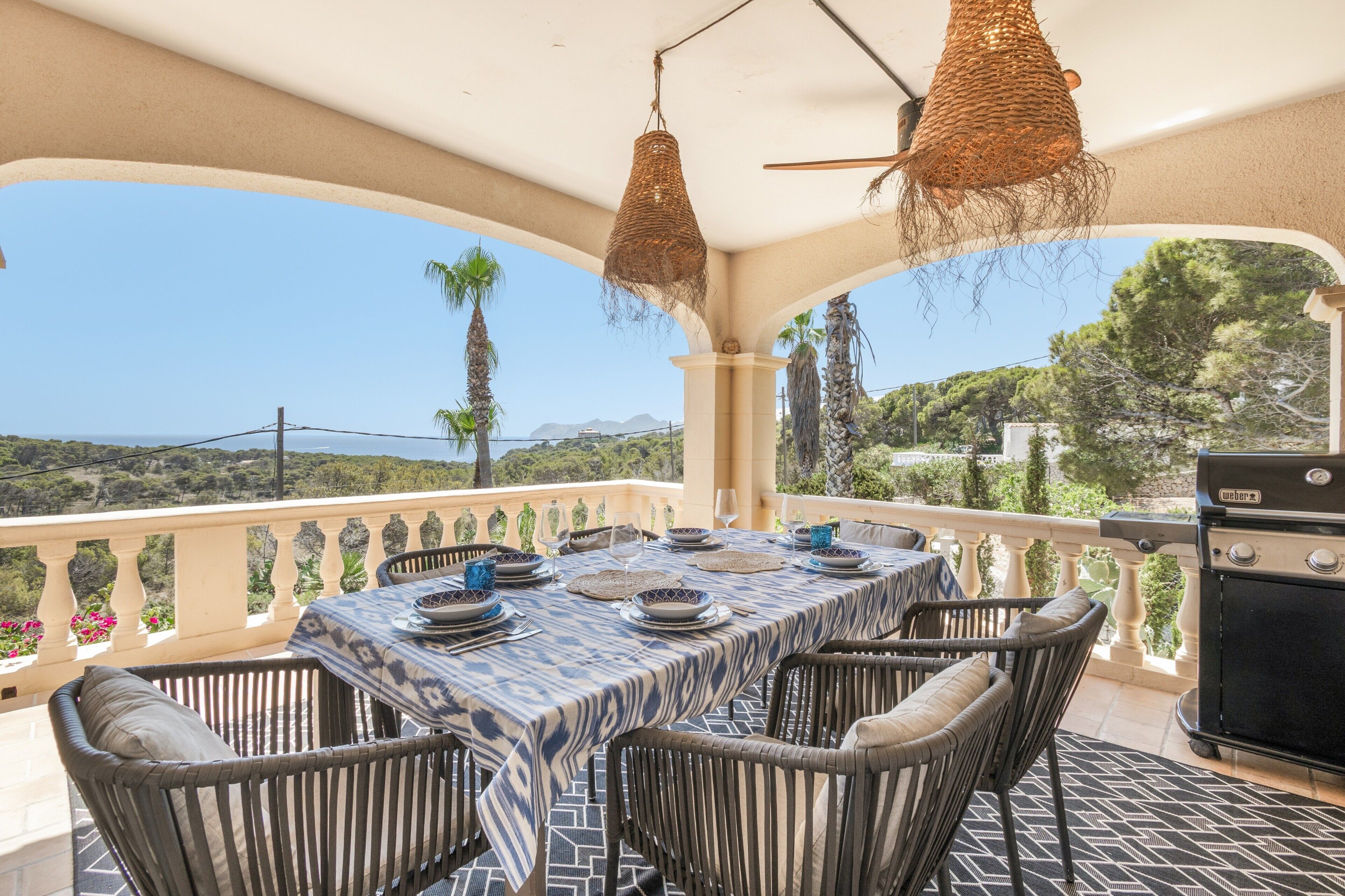 Un espace de restauration en plein air avec une table dressée pour quatre personnes, entourée de chaises en osier, sous une pergola agrémentée de guirlandes lumineuses.