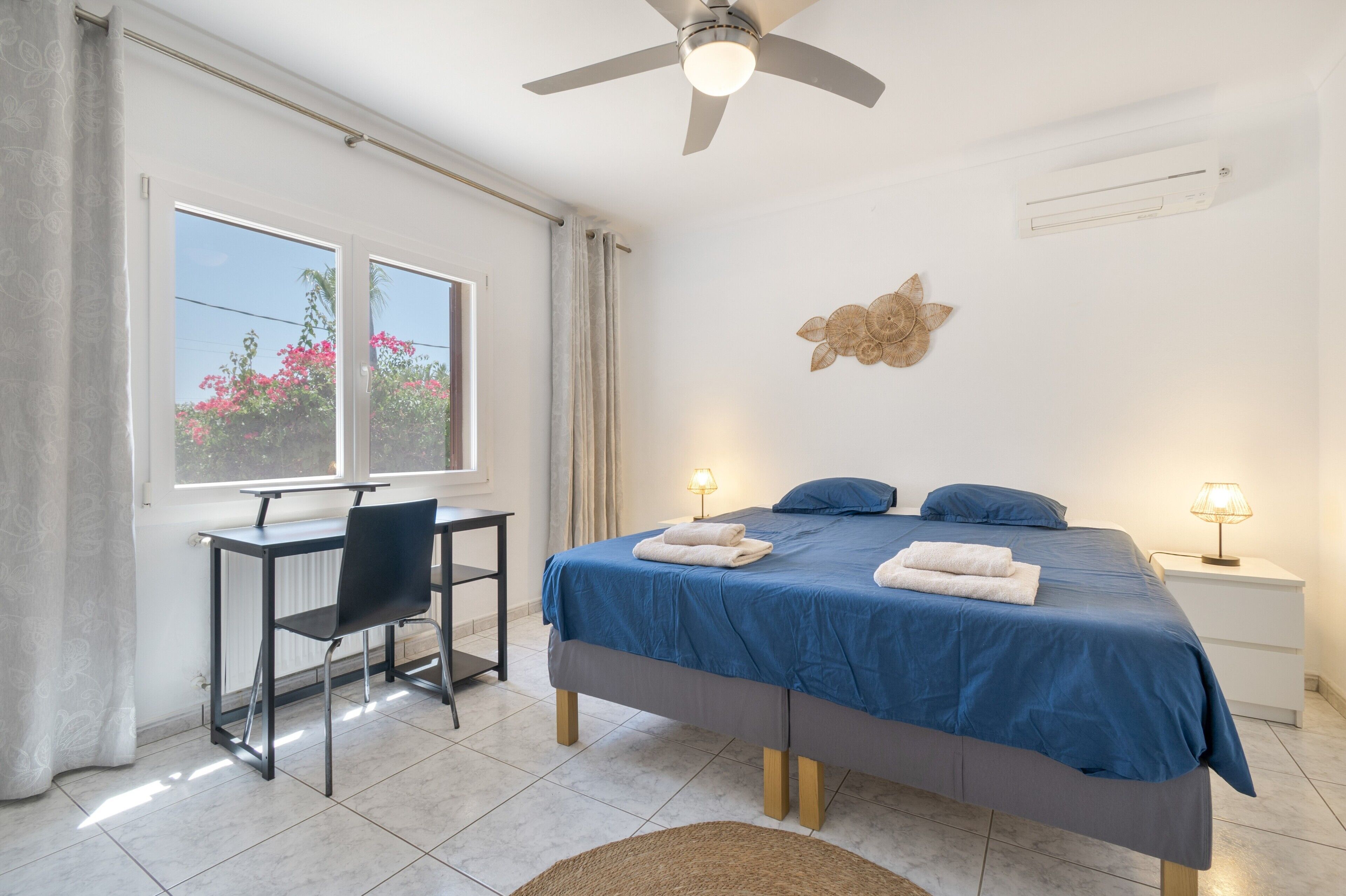 Un dormitorio con cama, escritorio, silla, ventilador de techo y ventana con cortinas.