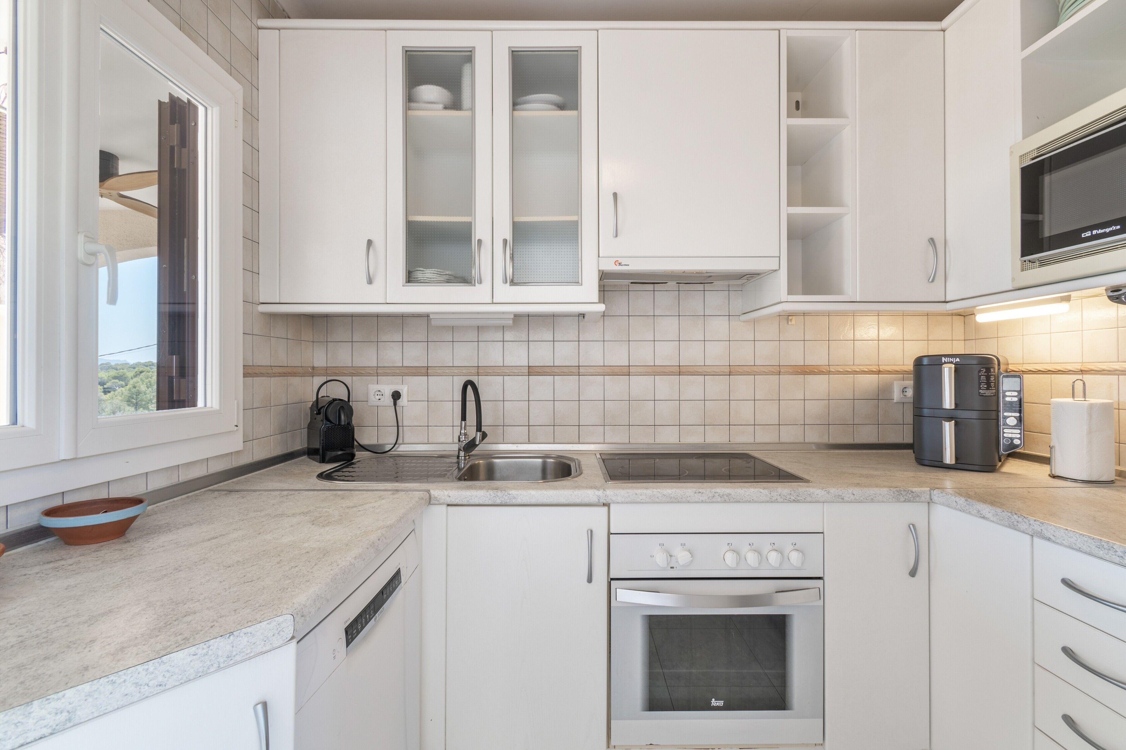 Una cocina moderna con gabinetes blancos, un lavamanos de acero inoxidable, horno empotrado y microondas.