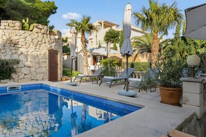 Una piscina con baldosas de piedra, sillas de descanso, sombrillas y plantas en macetas.