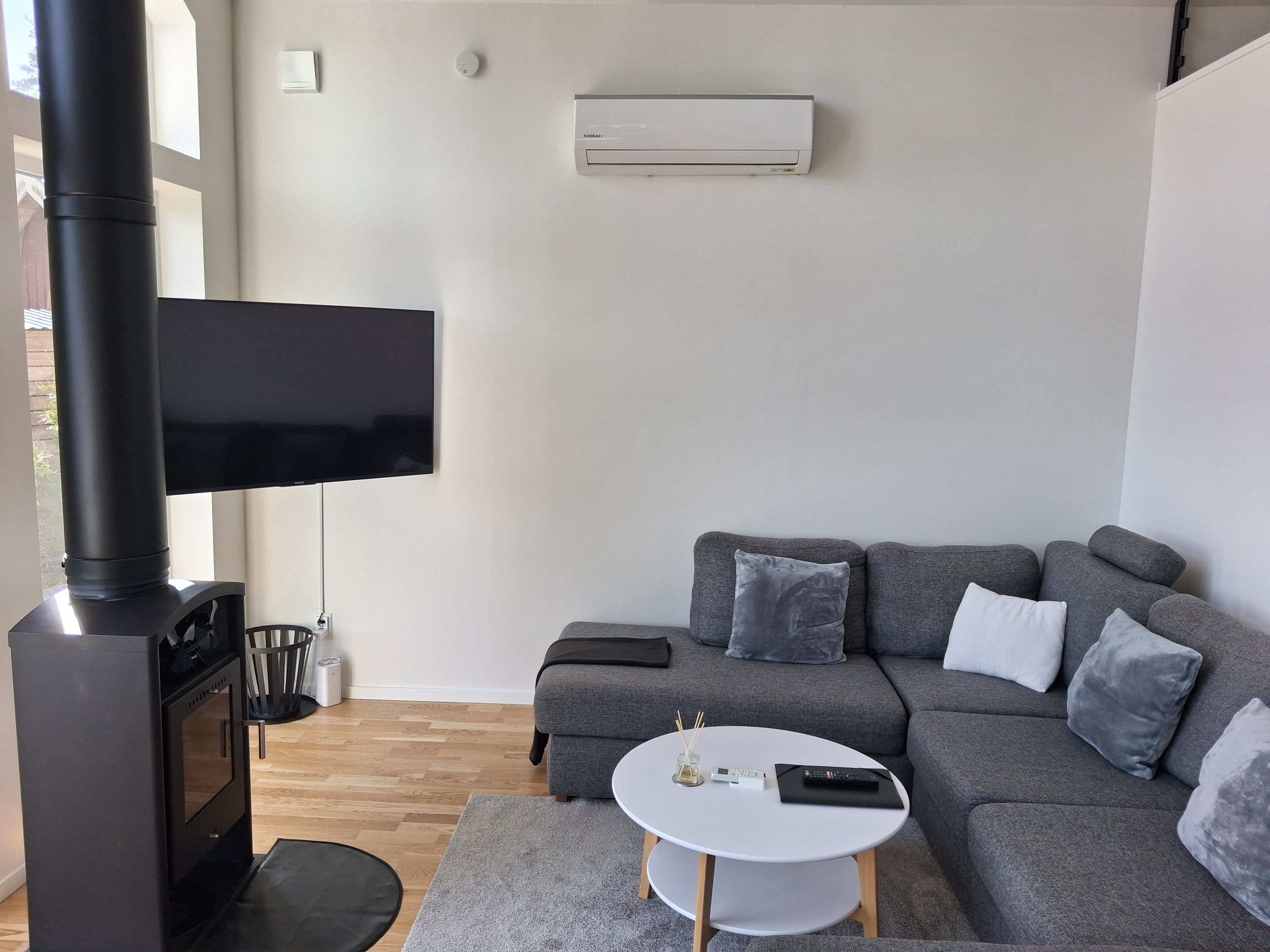 A modern living room with a fireplace, a flat-screen TV, a grey sofa with cushions, a white coffee table, and a small side table with a remote control.