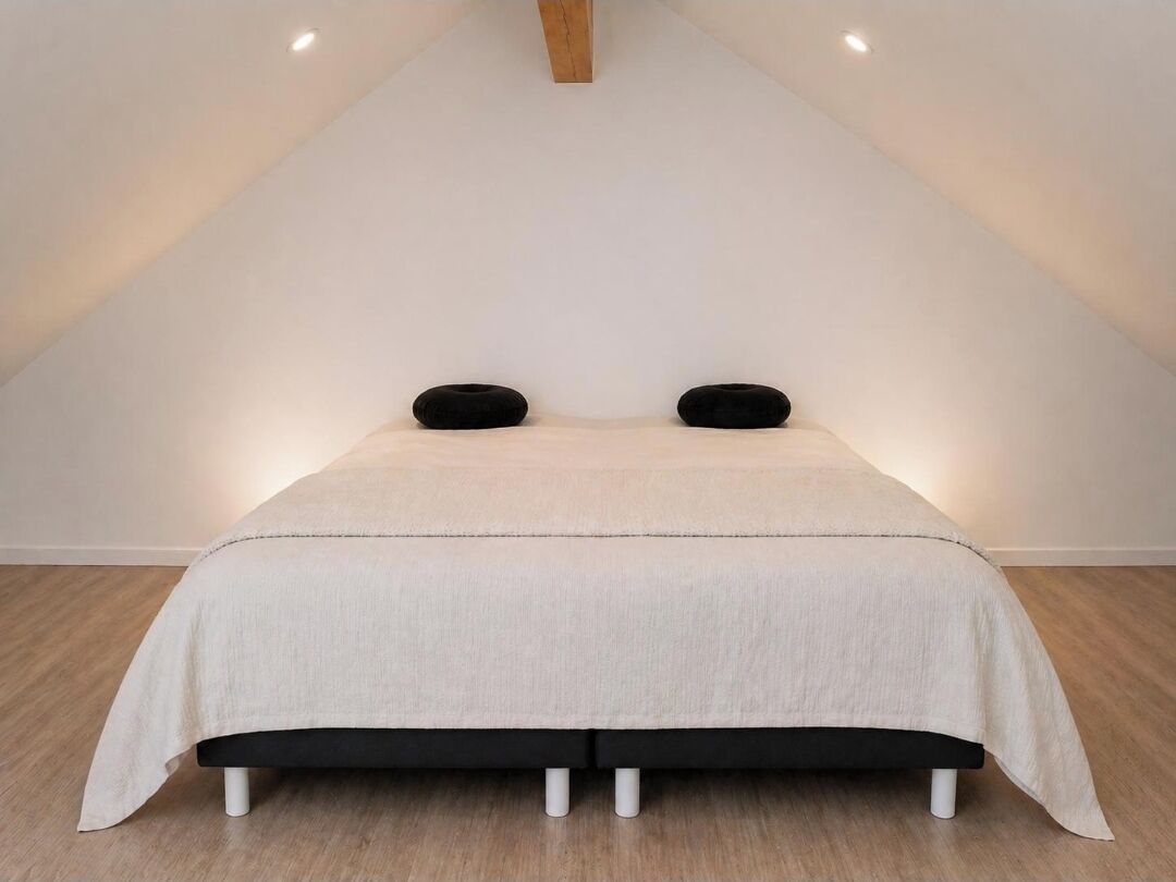 Una cama en habitación con techo inclinado, paredes blancas y suelo de madera.