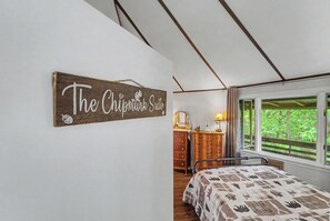 Ein Schlafzimmer mit Bett, Kommode, Spiegel und Fenster mit Blick auf Bäume.