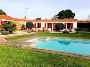 Ein Pool mit Holzterrasse, ein Steinhaus mit roten Fensterläden und einem gepflegten Rasen.