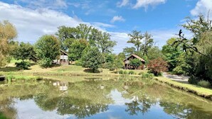 A tranquil pond with reflections, surrounded by lush greenery and two rustic cabins.