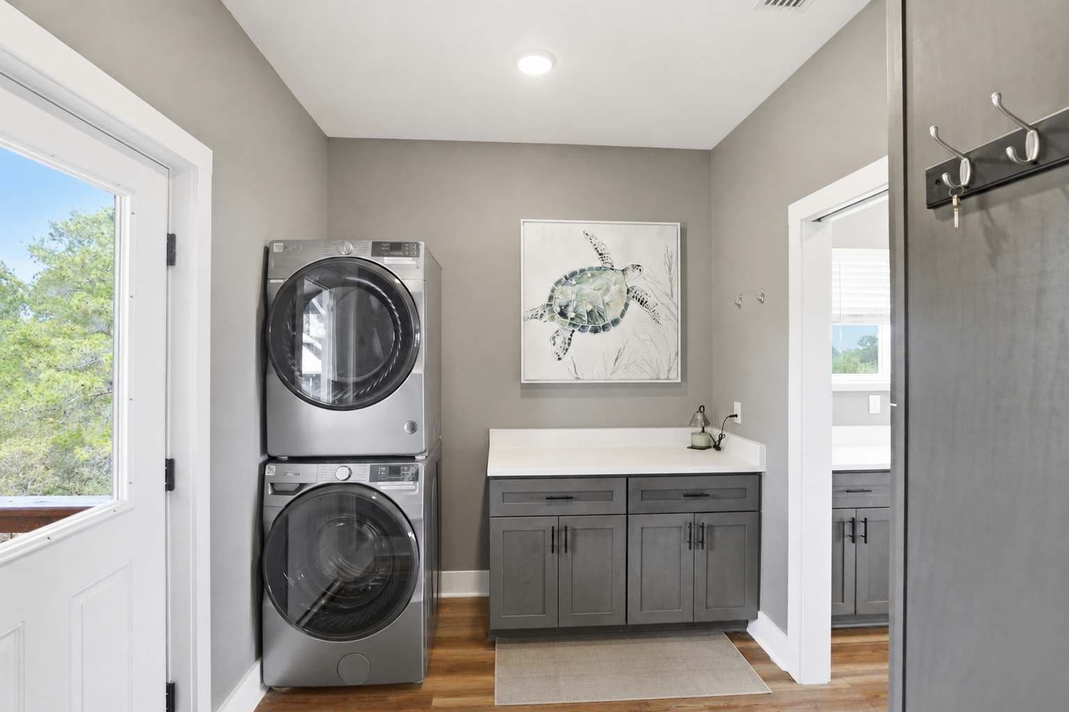 A laundry room with two stacked front-loading washing machines, a gray cabinet with drawers, a white countertop, and a wall-mounted artwork of a turtle.