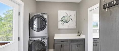 A laundry room with two stacked front-loading washing machines, a gray cabinet with drawers, a white countertop, and a wall-mounted artwork of a turtle.