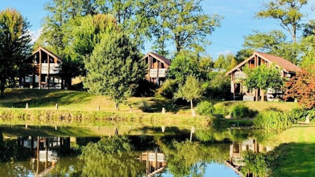 A lakeside property with wooden cabins, a pond, and lush greenery. - The Unusual Getaway • Nature Chalet & Hot Tub (Meilhac)
