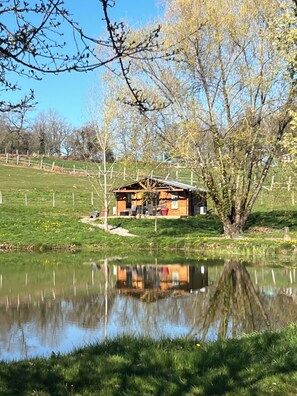 A wooden cabin with a porch, surrounded by a fenced field and a pond.