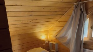 A wooden room with a bed, a bedside table, a wall-mounted lamp, and a window with curtains. - Les Chalets de Meilhac • Chalet N°2 (Meilhac)