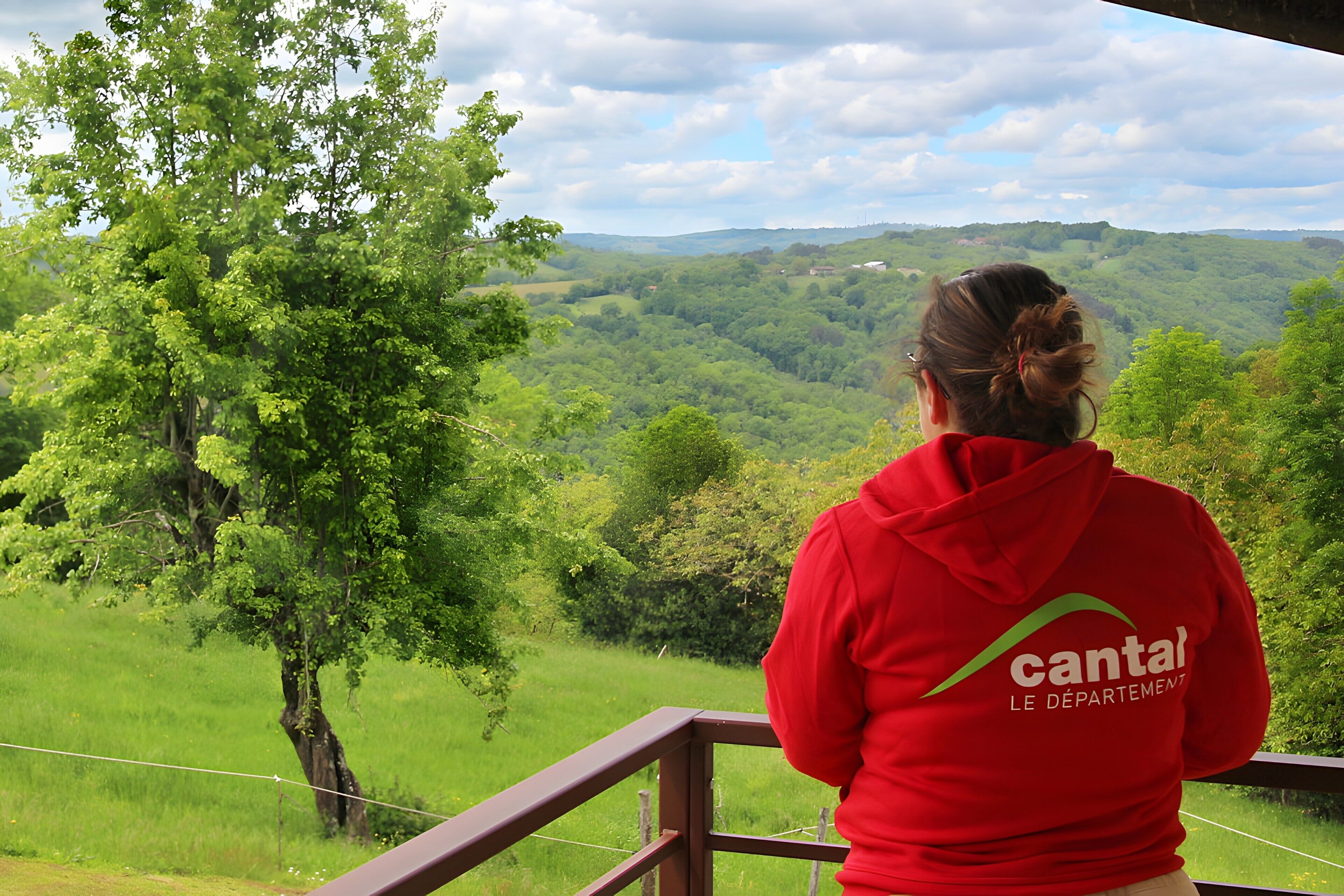 A person in a red hoodie with 'cantal' on the back, standing on a balcony with a view of a green landscape.