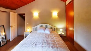 A bedroom with a sloped wooden ceiling, a bed with a white headboard, two bedside lamps, and a door leading to another room.