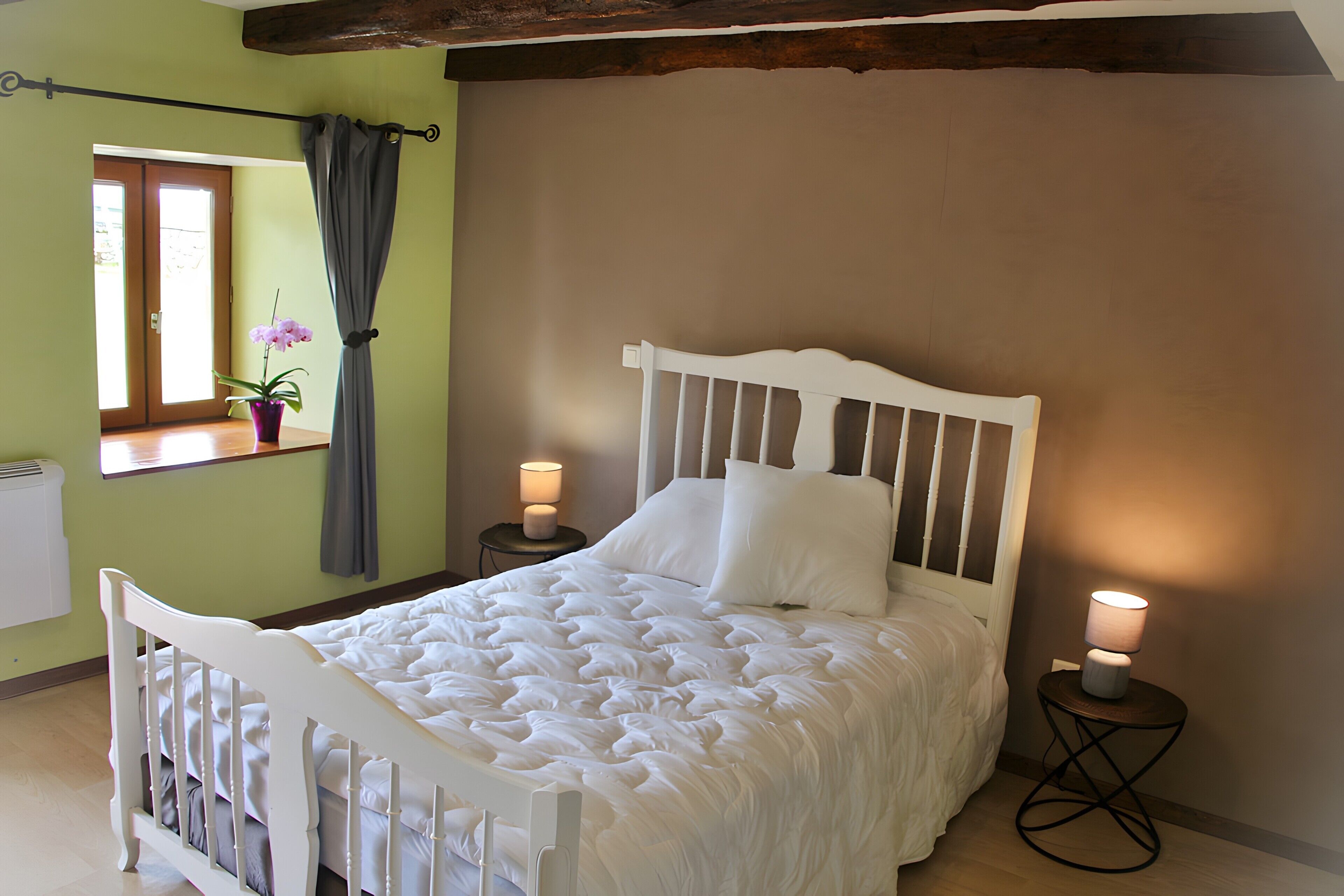 A bedroom with a white bed, a window with a curtain, a bedside lamp, and a radiator.