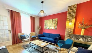 Un salon avec un canapé bleu, deux fauteuils en osier, une table basse en bois et un luminaire suspendu.