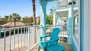 A turquoise balcony with a white railing, a blue Adirondack chair, and a view of the street and palm trees. - Mins from Beach • Sleeps 17 • Outdoor Seating (Destin)