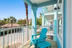A turquoise balcony with a white railing, a blue Adirondack chair, and a view of the street and palm trees. - Mins from Beach • Sleeps 17 • Outdoor Seating (Destin)