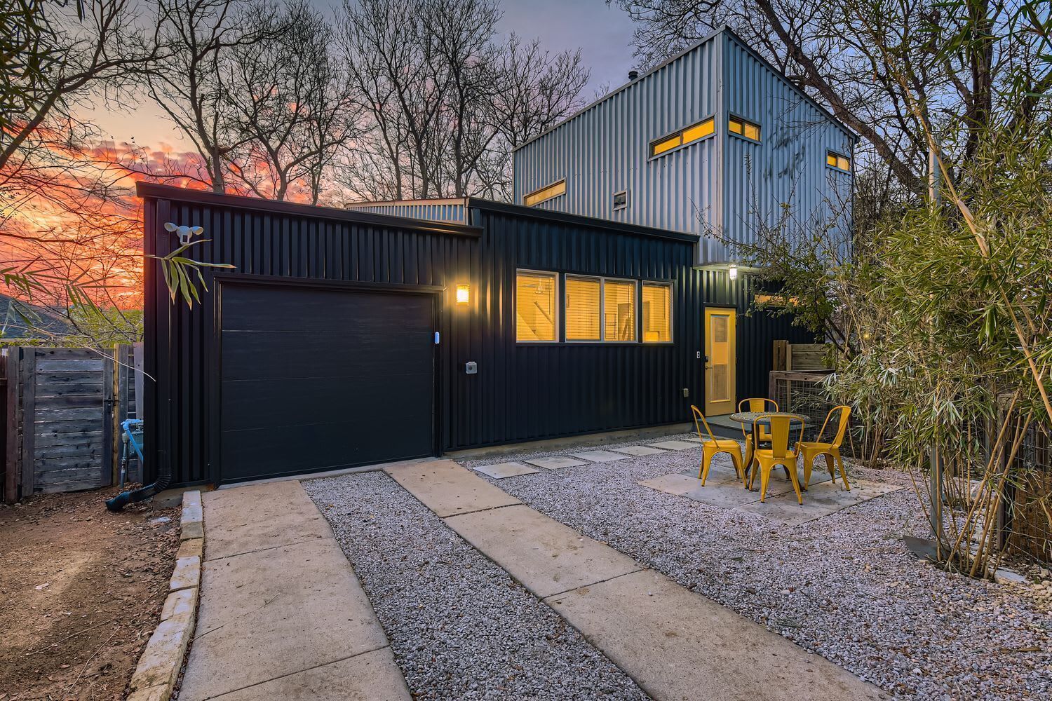 A modern house with a dark exterior, a garage, and a gravel driveway leading to a patio with yellow chairs.