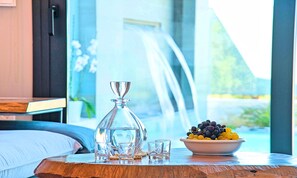 Une pièce comprenant un lit, une table avec un saladier de fruits, une carafe en verre et des verres.