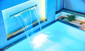 Une piscine extérieure moderne agrémentée d’une cascade, entourée d’un aménagement minimaliste et d’un petit jardin.