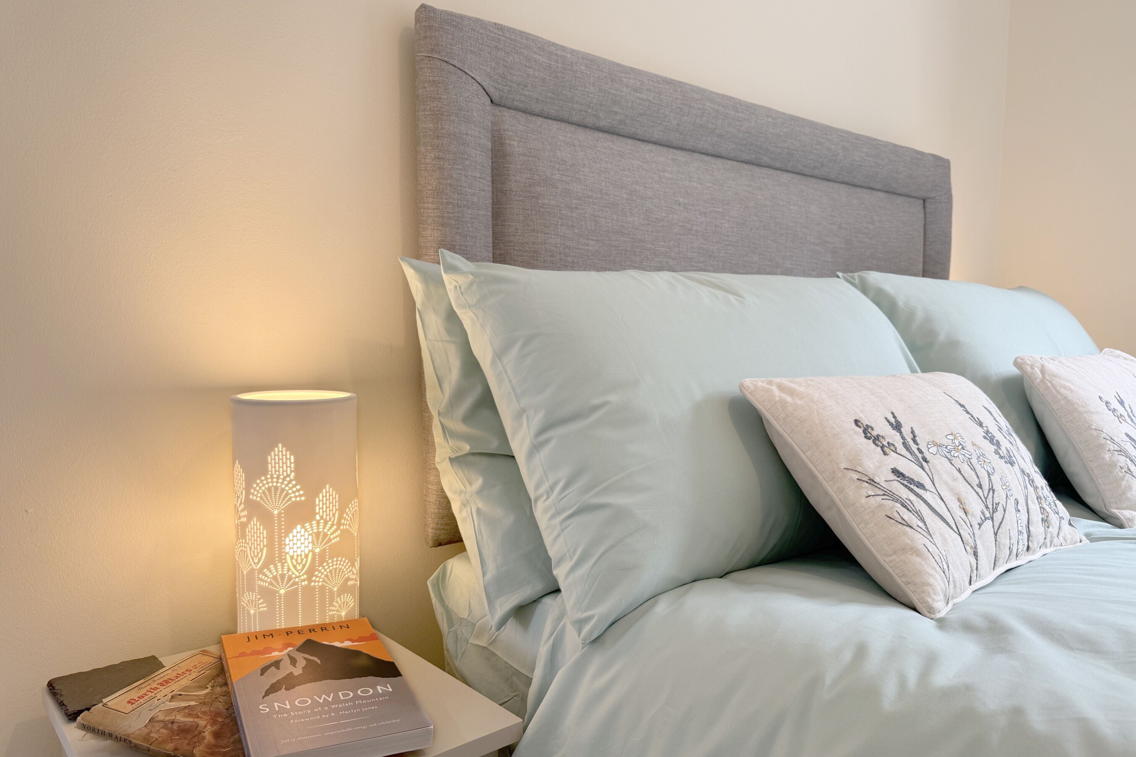 A bed with light blue bedding, a grey headboard, and decorative pillows with floral embroidery. A bedside table holds a book and a lamp with a leaf pattern.