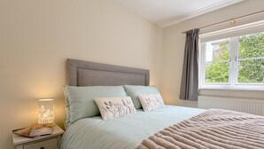 A bedroom with a bed, a nightstand, a lamp, and a window with curtains. - Muriau Tref - Three Bedroom Cottage, Sleeps 4 (Conwy)