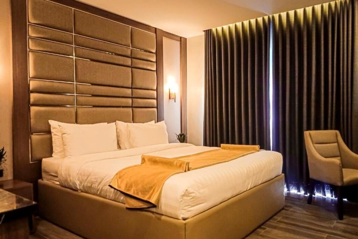 Ein modernes Hotelzimmer mit einem großen Bett, einem Kopfteil mit horizontalen Paneelen, einem Sessel und einem großen Fenster mit Vorhängen.