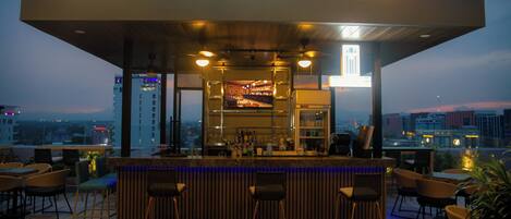 A rooftop bar with a bar counter, stools, and a city view.