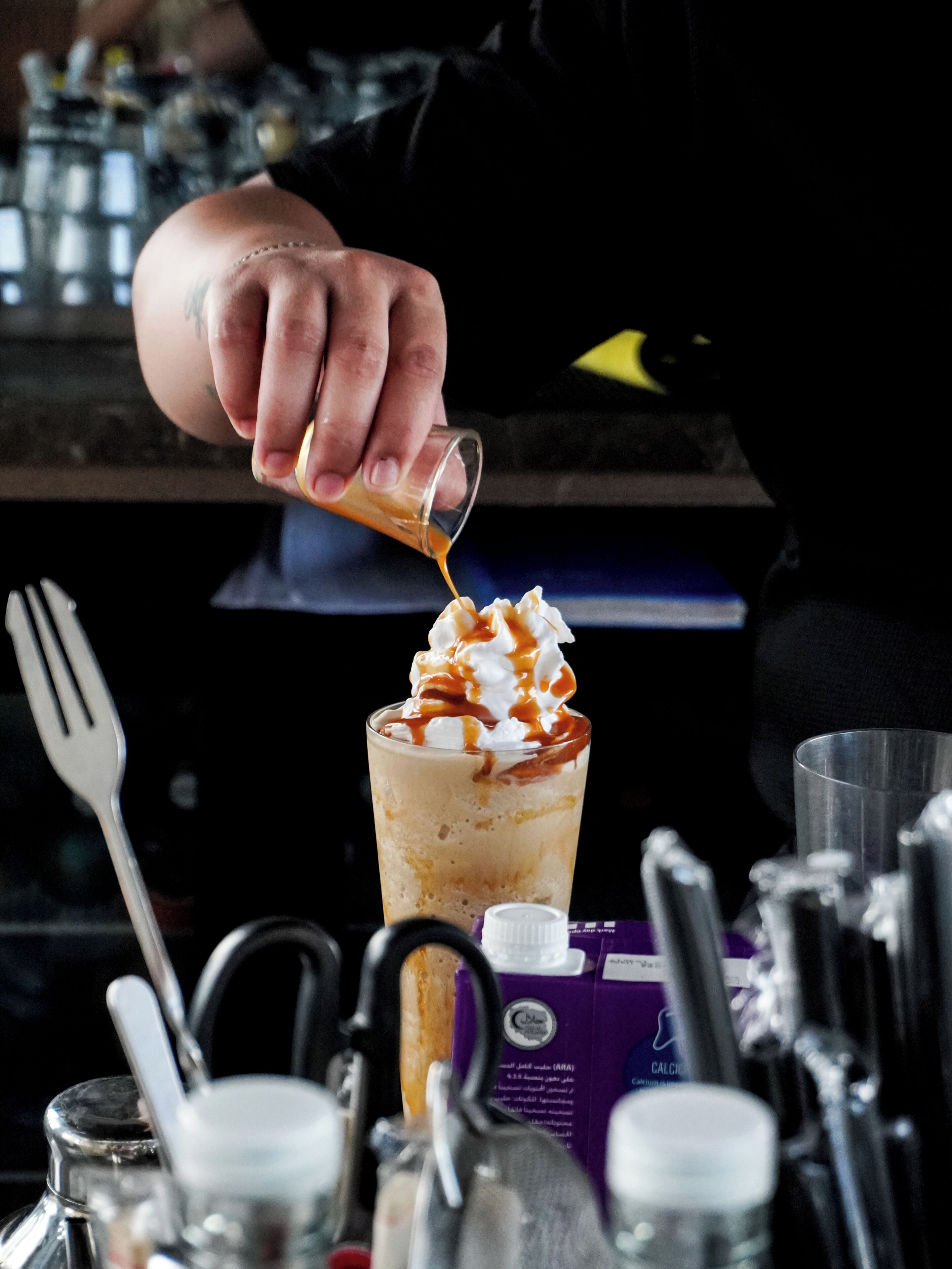 Une personne verse un liquide dans un verre garni de crème fouettée et nappé de caramel.