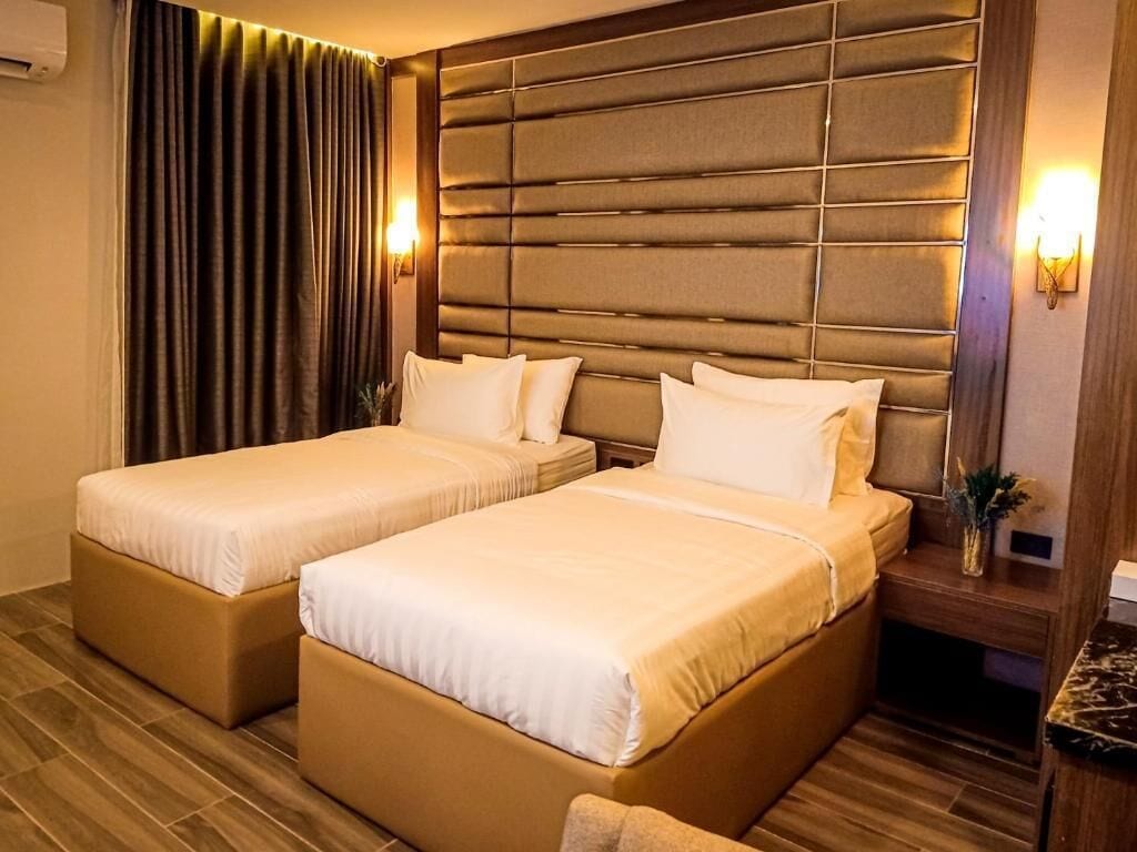A hotel room with two beds, a padded headboard, a nightstand, and a wall-mounted lamp.