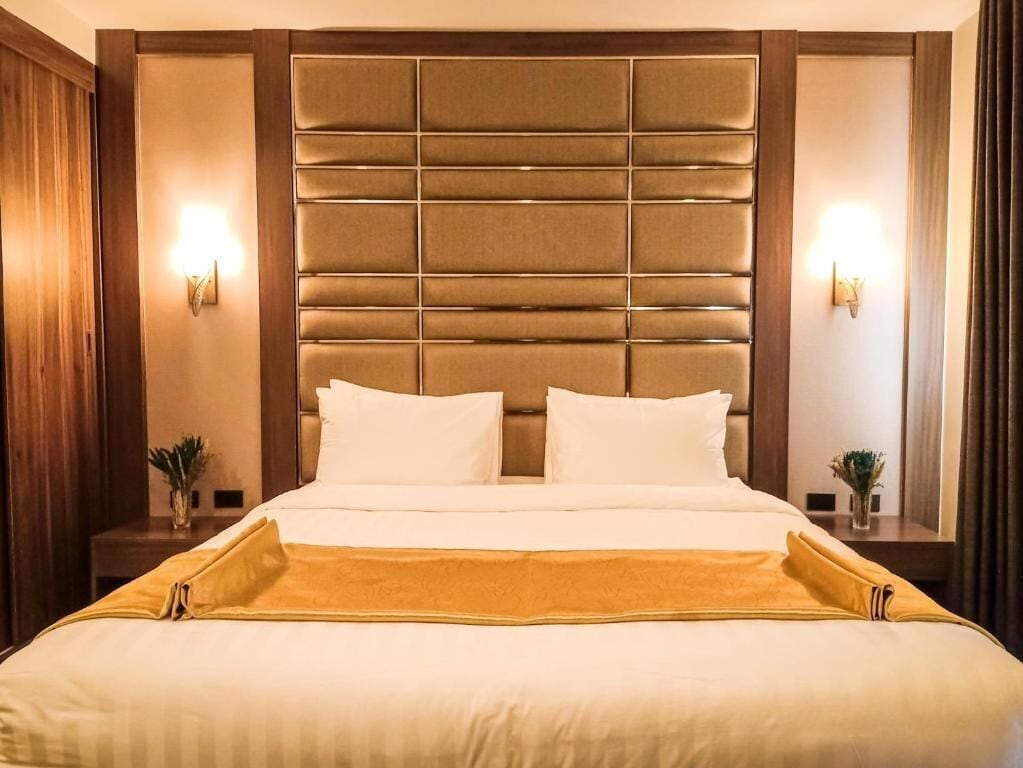 Habitación de hotel con una cama grande, cabecera acolchada, dos lámparas de noche y una mesita con una planta.
