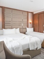 A hotel room with two beds, a wooden headboard, a wall-mounted light, and a brown armchair.