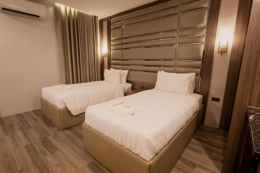 Une chambre d’hôtel avec deux lits, une grande tête de lit rembourrée, un climatiseur fixé au mur et un plancher en bois.