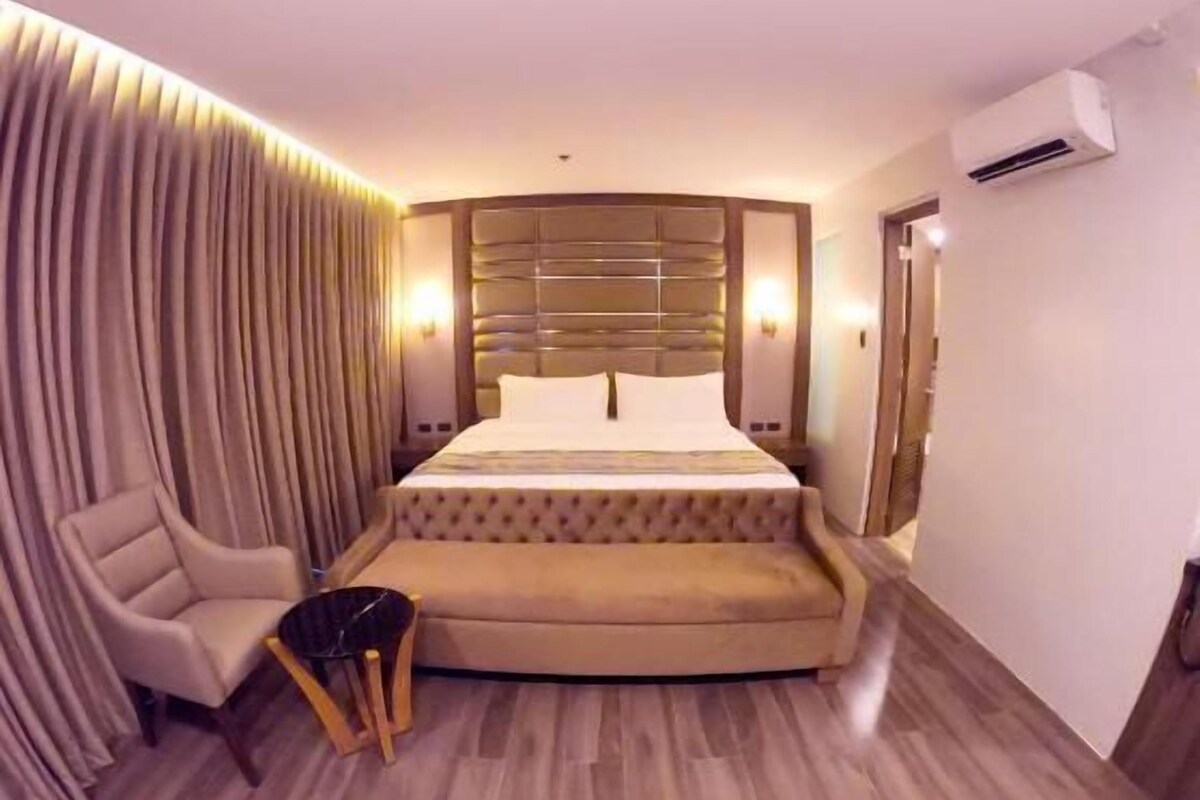 Ein Hotelzimmer mit einem großen Bett, einem Sessel, einem kleinen Tisch, einer Klimaanlage und einem Vorhang.