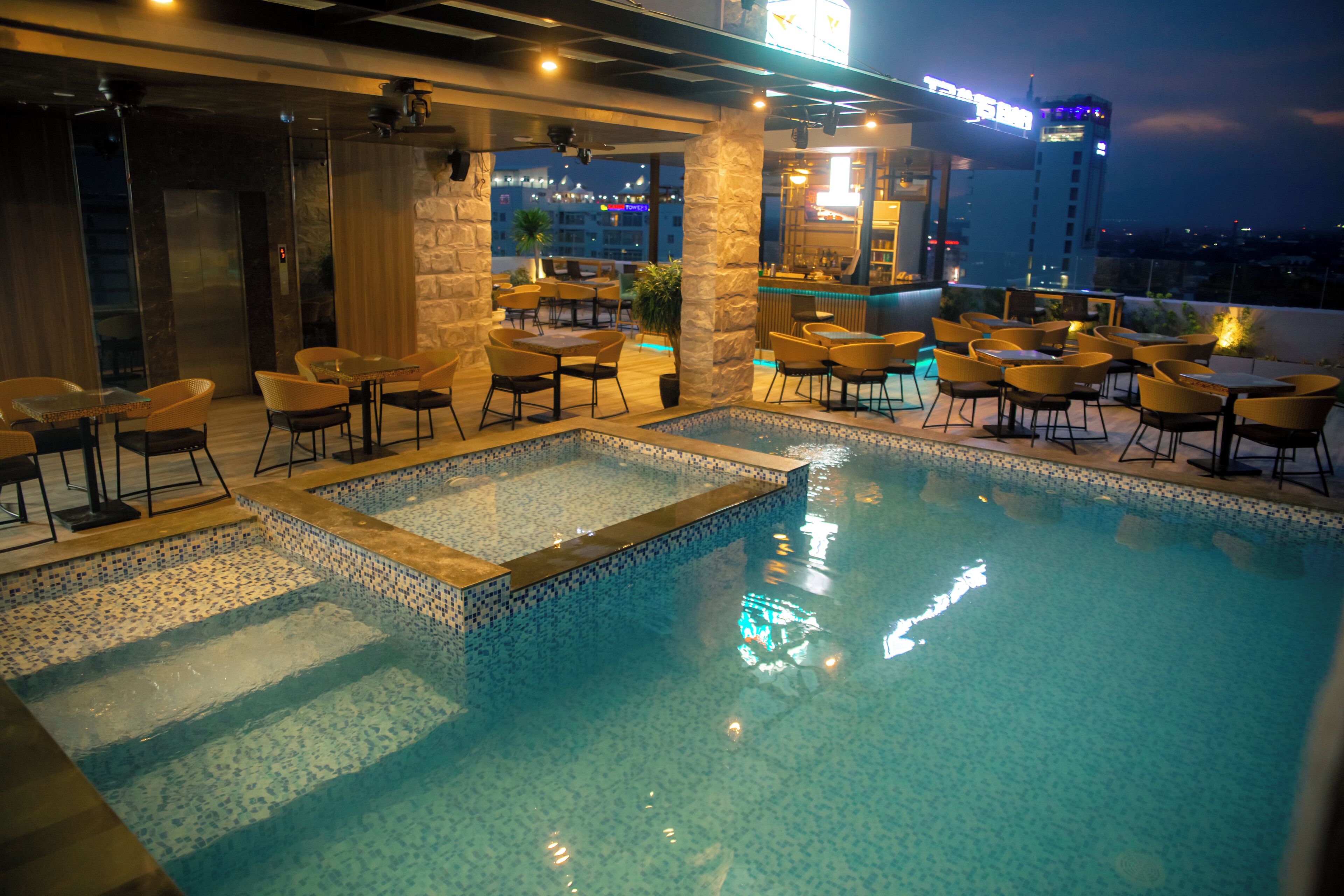 A rooftop pool with clear blue water, surrounded by yellow chairs and tables, and a bar area with a city view at night.
