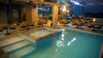 Una piscina en la azotea con agua azul cristalina, rodeada de sillas y mesas amarillas, y un área de bar con vista a la ciudad de noche.